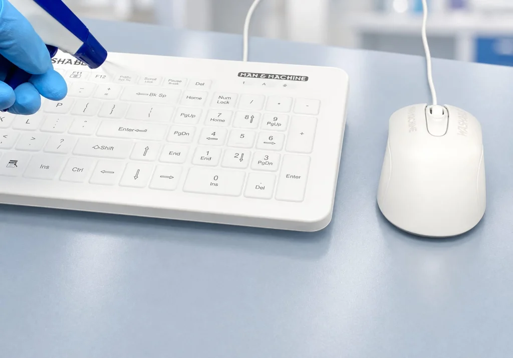 Gloved hand holding a spray nozzle over an MMI keyboard and mouse on a clinical worksurface